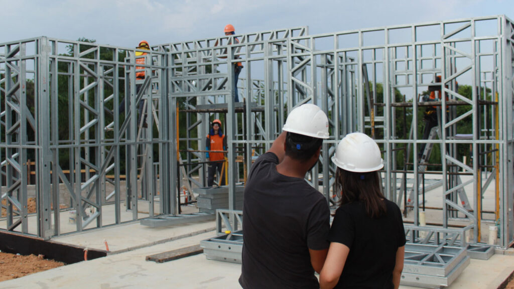 A foto mostra os fundadores da Vikka, Vitor e Karla, de costas. Eles olham para uma estrutura de uma residência em Light Steel Frame enquanto seus colaboradores realizam a obra.